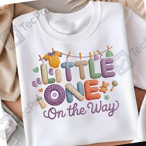 Puede incluir: Camiseta blanca con un diseño bordado colorido. El diseño incluye las palabras "LITTLE ONE" y "On the Way" en una fuente caprichosa, junto con ilustraciones temáticas de bebés como un body, botines y un osito de peluche.