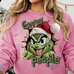 Könnte beinhalten: Rosa Sweatshirt mit einem grünen Grinch-Charakter, der einen Weihnachtsmannhut trägt und aus einem zerrissenen Wanddesign hervortritt. Der Text "Eww.. people" ist grün und weiß gestickt. Ein festliches und humorvolles Kleidungsstück.