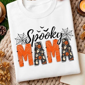 Könnte beinhalten: Ein weißes Sweatshirt mit Rundhalsausschnitt und dem schwarzen Schriftzug Spooky MAMA. Die Buchstaben sind mit orangefarbenen Spinnennetzen und einem schwarzen Hintergrund mit kleinen weißen Geistern, Kürbissen und Bonbons gefüllt.