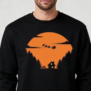May include: Black sweatshirt with a festive design. It features a silhouette of Santa's sleigh and reindeer against an orange sunset, with a house and trees silhouetted below, creating a Christmas scene.