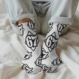 以下が含まれることがあります： 白い靴下には、星型のデザインの中に数字の「6」が入った黒いグラフィックパターンが繰り返されています。 靴下はリブ編みで、青いジーンズを履いた人が着用しています。