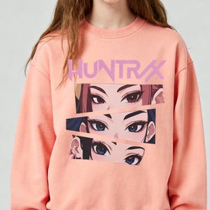 May include: A coral-colored sweatshirt featuring the word "HUNTRAX" in a light purple font. The graphic design includes three sets of stylized anime eyes in shades of brown and blue.