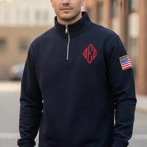 May include: Navy blue quarter-zip sweatshirt with a red diamond-shaped logo and an American flag patch on the sleeve. The sweatshirt has a high collar and a silver zipper. The logo appears to be an initial or monogram.