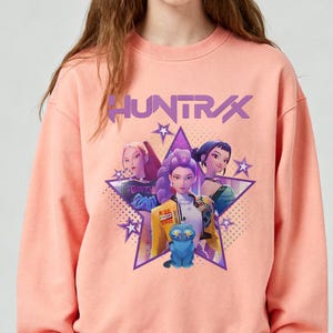 May include: A peach-colored sweatshirt featuring a graphic of three animated figures inside a purple star. The word "HUNTRAX" is printed above the star. The figures are wearing colorful outfits and have stylized hair.