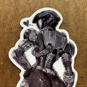 Op de afbeelding: Zwart-wit sticker met een gedetailleerde illustratie van een futuristisch gepantserde figuur, van achteren gezien. De figuur staat in een dynamische pose met een hartsymbool. De sticker heeft een witte rand.