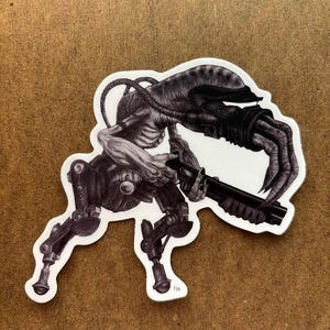 May include: Black and white sticker featuring a detailed illustration of a biomechanical creature holding a shotgun. The creature has a skeletal torso, mechanical legs, and a large, clawed hand. The sticker is die-cut with a white border.