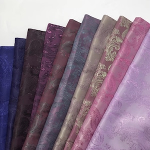 Puede incluir: Una variedad de muestras de tela en tonos morados, desde índigo oscuro hasta lavanda claro. Cada muestra presenta un estampado de cachemira, con diseños que varían en color y detalle. Las telas tienen un ligero brillo, lo que sugiere satén o seda.