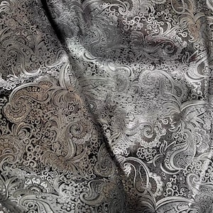 Puede incluir: Primer plano de una tela negra con un estampado de cachemira plateado. La tela está doblada, creando sombras y luces que enfatizan los intrincados diseños florales y arremolinados. El patrón es detallado y elegante.