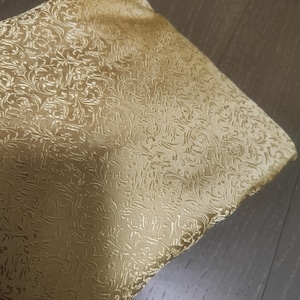 Peut inclure: Un morceau de tissu doré plié avec un motif floral en relief. Le tissu a un éclat subtil, et le motif est un motif répétitif de feuilles et de fleurs tourbillonnantes. La couleur est un or chaud et accueillant.