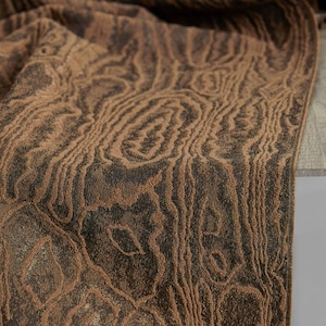 May include: Close-up of a brown and black fabric with a wood grain pattern. The fabric is folded, revealing the texture and color variations. The design features swirling lines and organic shapes.