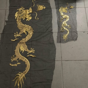 Könnte beinhalten: Zwei goldene Drachenapplikationen auf dunkelgrauem Stoff. Der größere Drache ist aufwendig detailliert, mit Schuppen und einem fließenden Körper. Der kleinere Drache ist eine vereinfachte Version. Beide sind auf einer gefliesten Oberfläche platziert.