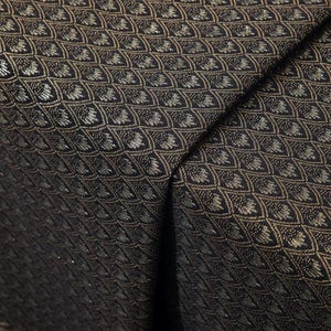 May include: Close-up of a folded fabric with a repeating pattern. The fabric is black with a gold and silver geometric design. The pattern is a series of interlocking shapes, creating a textured appearance. The fabric has a slight sheen.