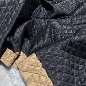 Puede incluir: Primer plano de una tela negra acolchada con un patrón de diamantes. La tela tiene una textura brillante y suave. La parte inferior es de color beige con un patrón de diamantes. Los bordes están acabados con un ribete decorativo.