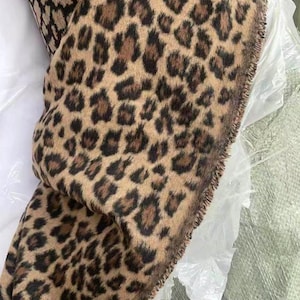 Könnte beinhalten: Ein gefaltetes Textil mit Leopardenmuster. Der Stoff hat einen beigefarbenen Hintergrund mit dunkelbraunen Flecken. Die Kanten des Stoffes haben eine Fransenborte. Das Textil wird wahrscheinlich für Kleidung oder Heimdekoration verwendet.