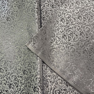 Puede incluir: Tela plateada con un patrón floral repetido. La tela tiene una apariencia texturizada y un brillo sutil. El diseño es de estilo damasco clásico, adecuado para diversas aplicaciones decorativas.