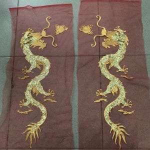 May include: Two gold and silver dragon appliques on a maroon mesh background. The dragons are intricately detailed with scales and flowing designs. Each dragon has a curved body and is facing the opposite direction.