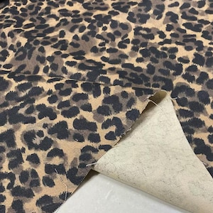 Peut inclure: Gros plan sur un tissu à imprimé léopard. Le motif présente des taches marron foncé et noires sur un fond beige. Une section du tissu est pliée, révélant un envers beige. Le tissu semble être de poids moyen.