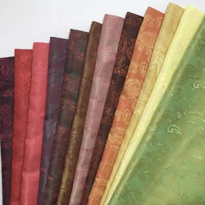Könnte beinhalten: Eine Auswahl an Stoffen mit Paisley-Muster in verschiedenen Farben, darunter tiefes Burgunderrot, Rosa und Olivgrün. Der Stoff hat einen dezenten Glanz, und das Paisley-Design ist sichtbar.