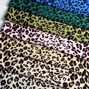 May include: Assortment of leopard print fabric swatches in various colors. The fabrics include blue, green, yellow, pink, and tan, each with black and brown spots. The swatches are arranged in a layered, diagonal pattern.