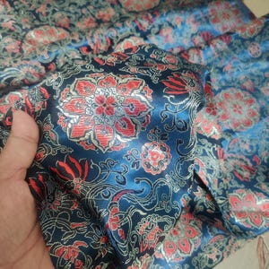 May include: A close-up of a dark blue fabric with a floral pattern in red and silver. The fabric is folded, revealing its texture and design. The floral pattern is intricate and detailed, with a satin sheen.