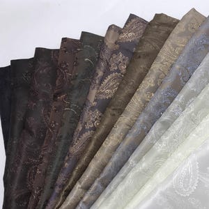 Puede incluir: Una variedad de muestras de tela con estampado de cachemira en varios colores, incluyendo negro, marrón, gris y crema. La tela tiene un brillo sutil, y el diseño de cachemira es visible en las muestras.