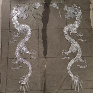 May include: Two silver dragon appliques with intricate detailing, including scales, claws, and flowing tails. The dragons are positioned on a textured, gray surface, with additional decorative elements. The appliques are likely for fabric embellishment or crafting projects.
