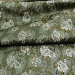 May include: A sage green fabric featuring a repeating pattern of white floral blossoms and intricate gold paisley swirls. The textile has a textured appearance with soft folds creating depth across the surface of the patterned material.