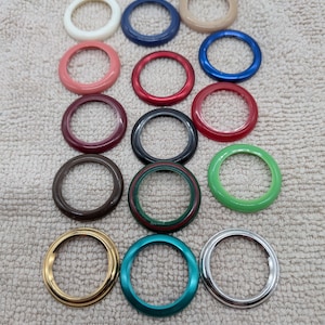 Puede incluir: Una colección de objetos circulares coloridos en varios tonos, incluyendo blanco, azul, rojo y verde. Los objetos parecen ser anillos decorativos o componentes, posiblemente para joyería o manualidades. La disposición está sobre una superficie neutra y texturizada.