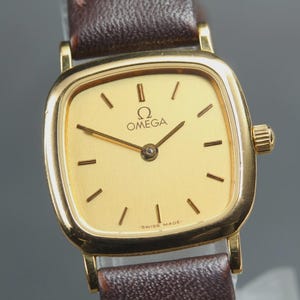 May include: A gold-tone Omega wristwatch with a square-shaped case featuring rounded corners and a gold dial. The watch has thin black hour markers, gold hands, and a dark brown leather strap. The text OMEGA and SWISS MADE are visible.