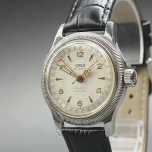 Puede incluir: Un reloj de pulsera automático Oris con caja de acero inoxidable plateado, esfera color crema texturizada y manecillas doradas. El reloj cuenta con una manecilla de fecha, números arábigos y una correa de cuero negro con relieve.