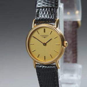 Peut inclure: Une montre-bracelet analogique dorée dotée d'un boîtier rond avec un cadran doré, des aiguilles noires fines et des index noirs. Le cadran affiche le logo Longines et le mot Swiss en bas. Elle possède un bracelet en cuir noir texturé.