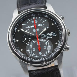 May include: A Seiko 5 chronograph watch with a black dial, silver-tone case, and black textured leather strap. The dial features a 1/20 sec sub-dial, a date window, and a red central second hand. The text reads Seiko 5 Chronograph 100M.