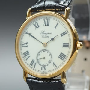 May include: A gold-toned Longines Quartz watch with a cream-colored face and black Roman numerals. The watch features a small seconds dial and black leather band. The watch is a classic timepiece.