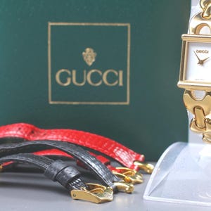 Puede incluir: Un reloj Gucci dorado con esfera blanca y correa de eslabones dorados a juego. El reloj se exhibe sobre un soporte transparente, con correas de reloj adicionales de cuero rojo y negro. Una caja verde con el logotipo de Gucci está en el fondo.