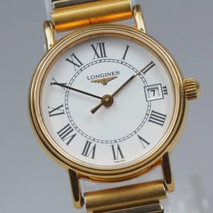 May include: A gold-tone wristwatch with a round case and a white dial featuring black Roman numeral hour markers. The dial displays the Longines logo and name below the twelve o'clock position, with a date window at the three o'clock position.