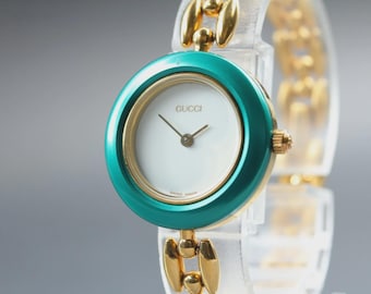 May include: A Gucci wristwatch featuring a round white dial with gold-tone hands and a teal bezel. The watch case is gold-tone, and the bracelet consists of alternating gold-tone metal links and clear, translucent acrylic links, creating a distinct contrast.