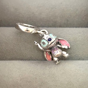 May include: A silver charm featuring a cartoon character with blue, pink, and purple enamel accents. The charm has a loop for attaching to a bracelet or necklace. The character has large ears and a playful pose.