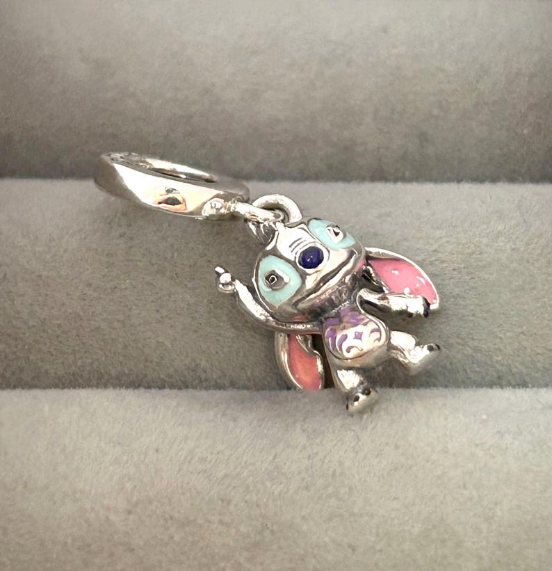 May include: A silver charm featuring a cartoon character with blue, pink, and purple enamel accents. The charm has a loop for attaching to a bracelet or necklace. The character has large ears and a playful pose.