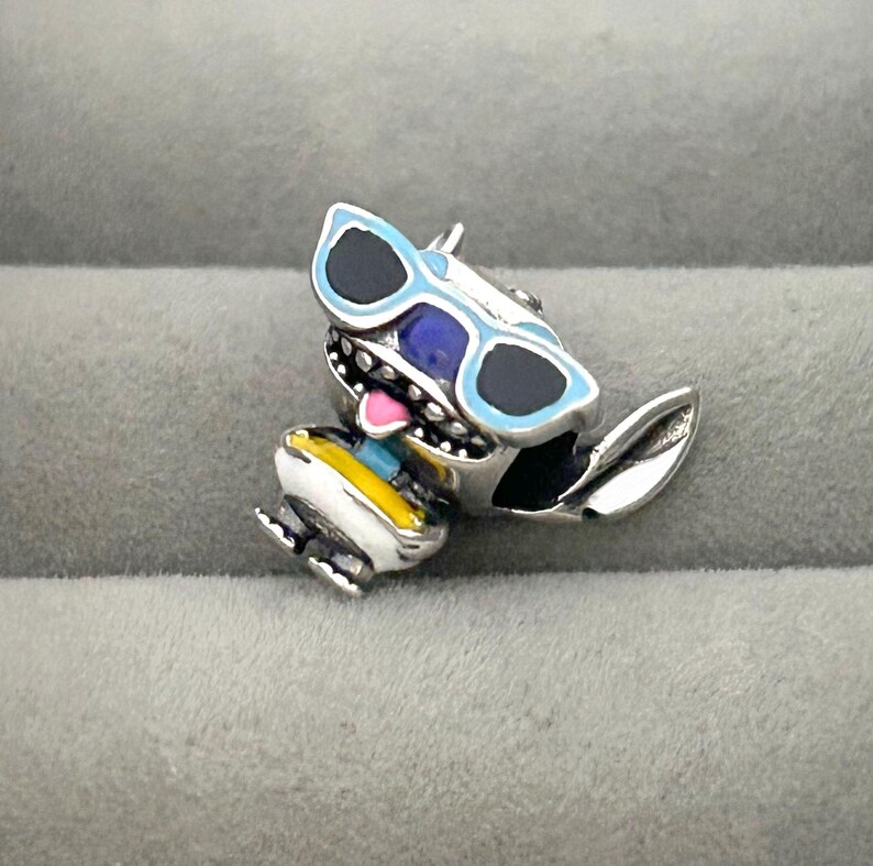 K&ouml;nnte beinhalten: Ein silberner Charm-Anh&auml;nger mit einer Comicfigur mit blauer Sonnenbrille und bunter Kleidung. Der Charm hat schwarze, blaue, gelbe und wei&szlig;e Akzente. Die Figur l&auml;chelt und hat eine rosa Zunge.