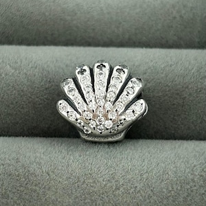 以下が含まれることがあります： 透明なクリスタルが輝く、シルバーの貝殻チャーム。チャームは扇形で、貝殻の各部分には複数のクリスタルが飾られています。クリスタルは光を捉え、鮮やかな効果を生み出します。このジュエリーはペンダントまたはチャームである可能性があります。