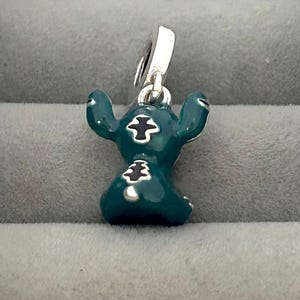 May include: A teal-colored charm featuring a cartoon character with large ears and black markings. The charm has a silver-colored loop for attaching to a bracelet or necklace. The character is in a sitting position.