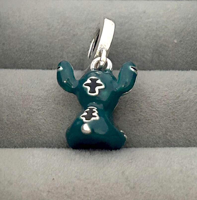 May include: A teal-colored charm featuring a cartoon character with large ears and black markings. The charm has a silver-colored loop for attaching to a bracelet or necklace. The character is in a sitting position.