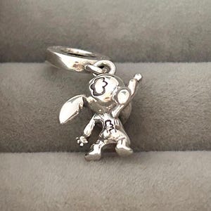 May include: A silver charm depicting a cartoon character with large ears, a round head, and a raised arm. The charm has a loop for attaching to a bracelet or necklace. The character's features are detailed with etched lines.