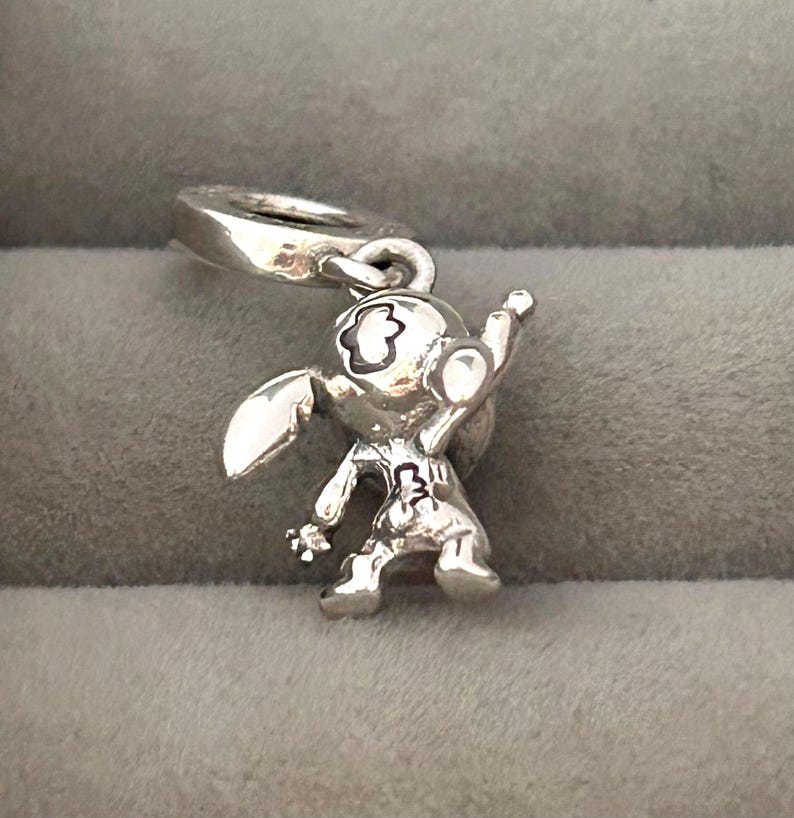 May include: A silver charm depicting a cartoon character with large ears, a round head, and a raised arm. The charm has a loop for attaching to a bracelet or necklace. The character's features are detailed with etched lines.