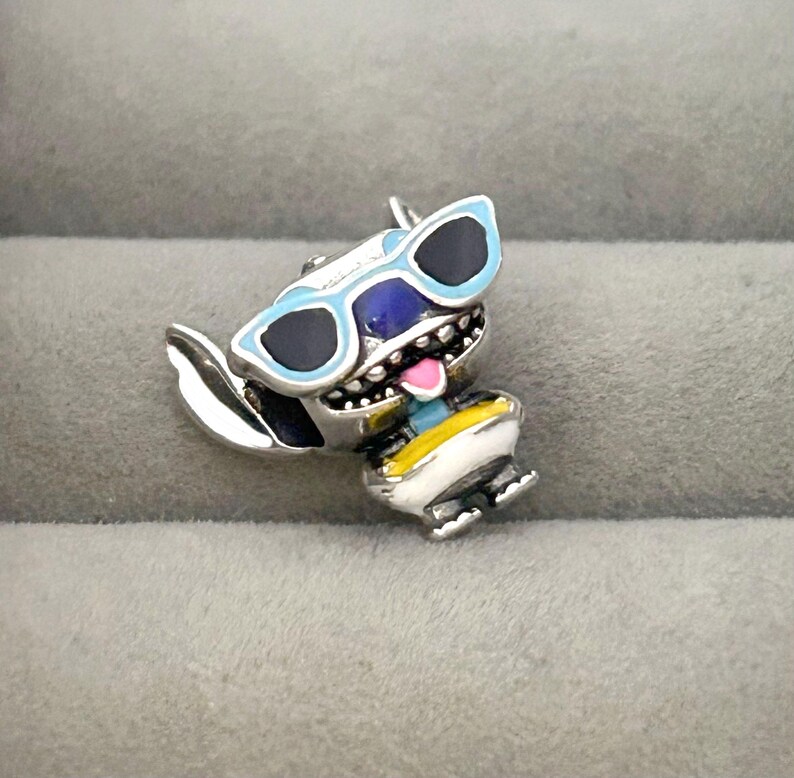K&ouml;nnte beinhalten: Ein silberner Charm-Anh&auml;nger mit einer Zeichentrickfigur mit blauer Sonnenbrille, blauem Gesicht und rosa Zunge. Die Figur tr&auml;gt ein gelb-blaues Outfit. Der Charm-Anh&auml;nger ist vor einem grauen Hintergrund platziert.