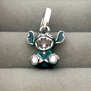 May include: A teal and silver charm featuring the character Stitch from Lilo & Stitch. The charm has a detailed design with Stitch's signature large ears, open mouth, and small feet. The charm is suspended from a silver loop.