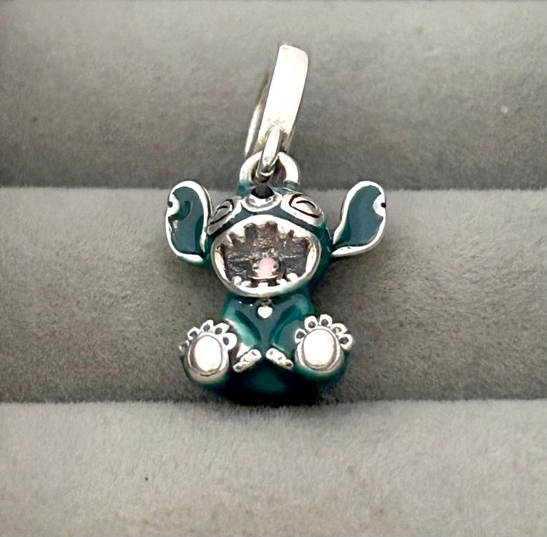 May include: A teal and silver charm featuring the character Stitch from Lilo & Stitch. The charm has a detailed design with Stitch's signature large ears, open mouth, and small feet. The charm is suspended from a silver loop.