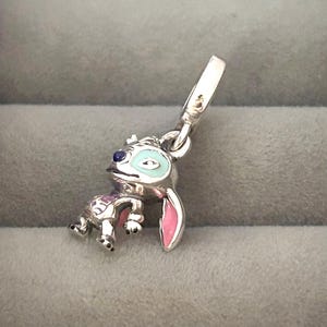 May include: A silver charm featuring the character Stitch from Lilo & Stitch. The charm has blue, pink, and white enamel details. The charm is in a crouching position with a loop for attaching to a bracelet or necklace.