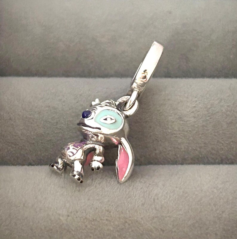 May include: A silver charm featuring the character Stitch from Lilo & Stitch. The charm has blue, pink, and white enamel details. The charm is in a crouching position with a loop for attaching to a bracelet or necklace.