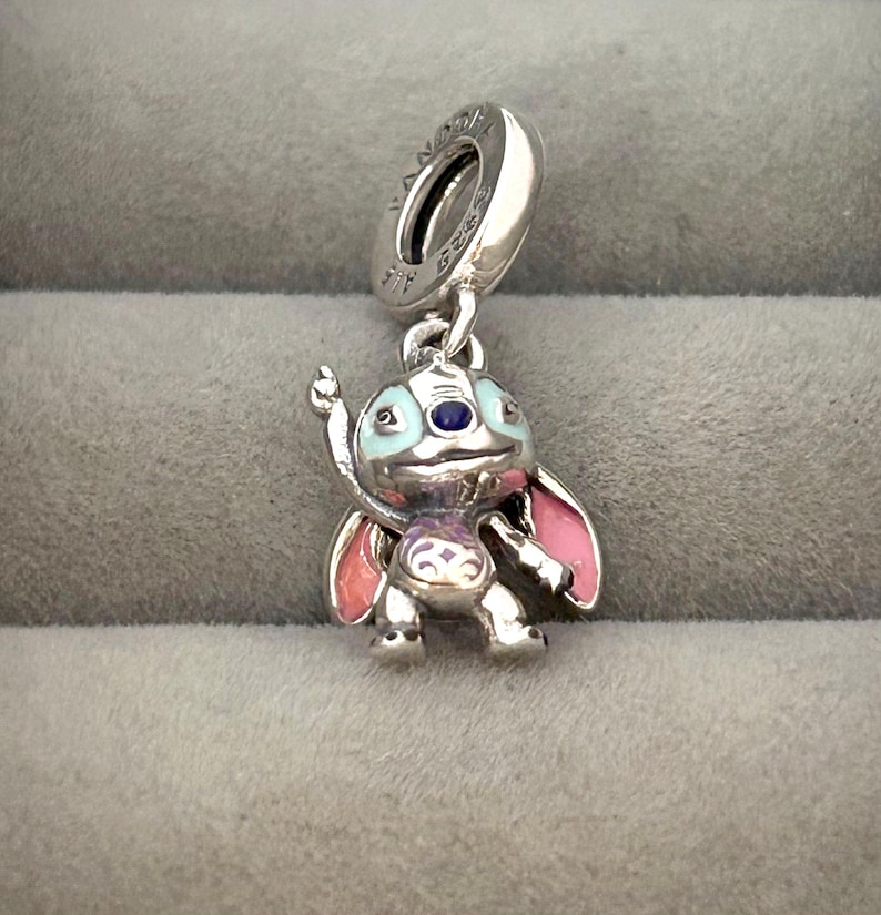 May include: A silver charm featuring a cartoon character with blue, pink, and purple enamel accents. The charm has a loop at the top for attaching to a bracelet or necklace. The character has large ears and is in a standing pose.